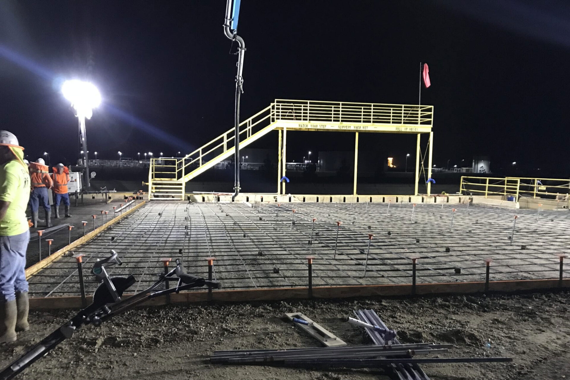 Nighttime construction site with a focus on a newly laid concrete foundation reinforced with a grid of steel rebar. Workers in high-visibility clothing and hard hats are present, with a portable light tower illuminating the area. In the background, construction equipment and industrial structures are faintly visible under the dark sky.
